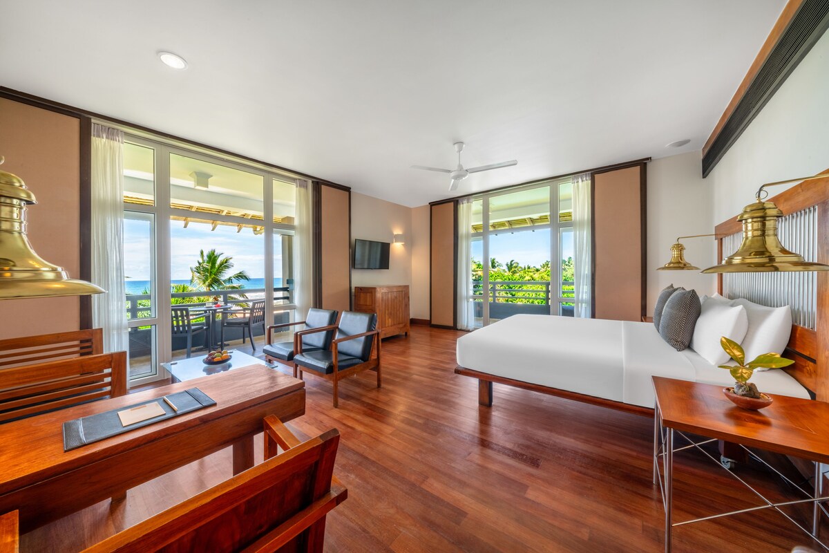 Quarto de hotel moderno com uma cama grande, móveis de madeira, área de refeições e varanda com vista para o oceano.