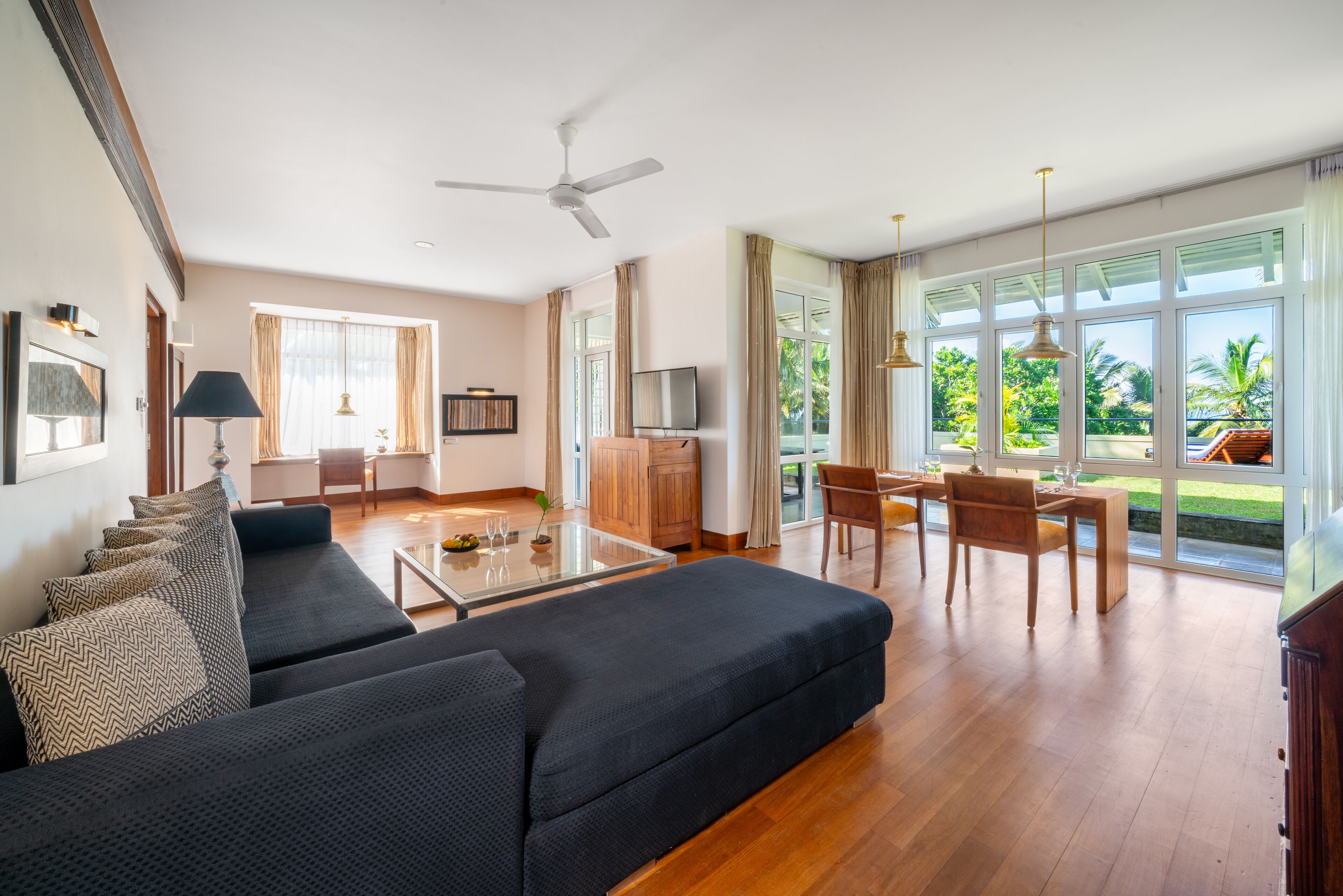 A spacious living room with a dark-colored sofa, a glass coffee table, and a dining area with wooden furniture.