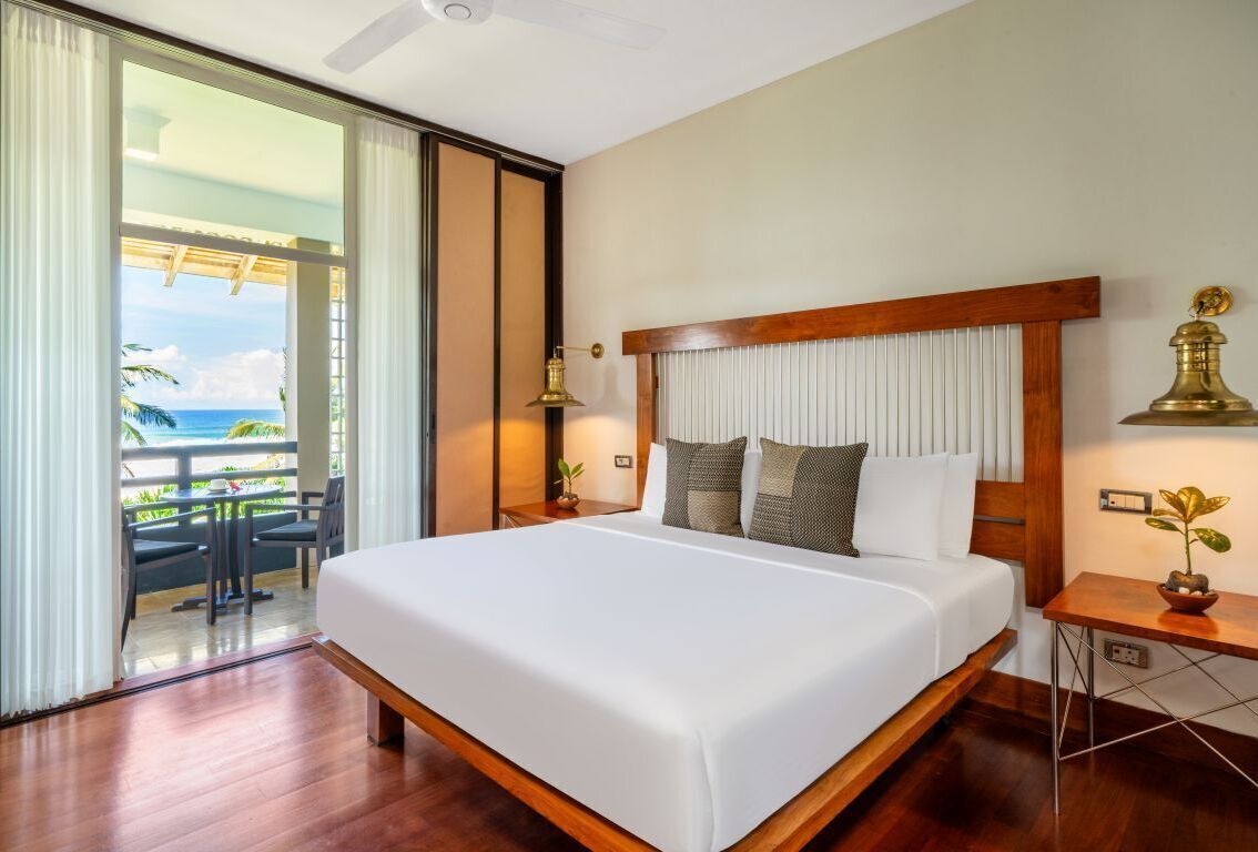 Une chambre d’hôtel avec un grand lit, une vue sur l’océan, un balcon avec des sièges d’extérieur et une lampe en laiton.