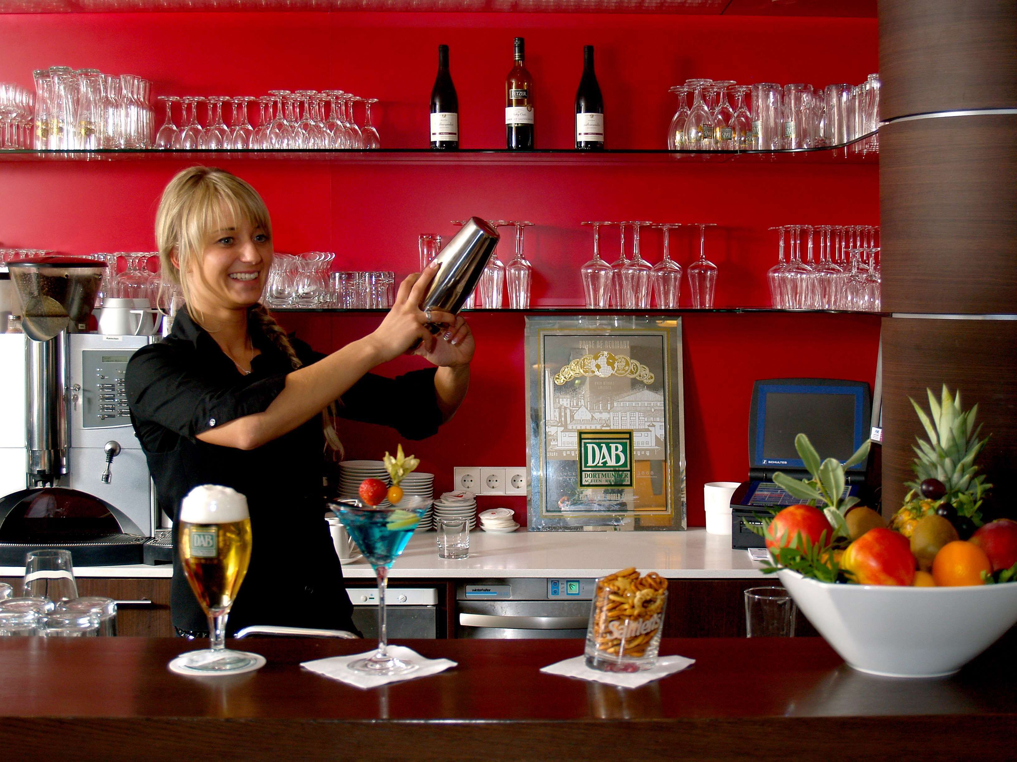 Ein Barkeeper bereitet ein Getränk zu, im Hintergrund sind Regale mit Weingläsern und Flaschen zu sehen.