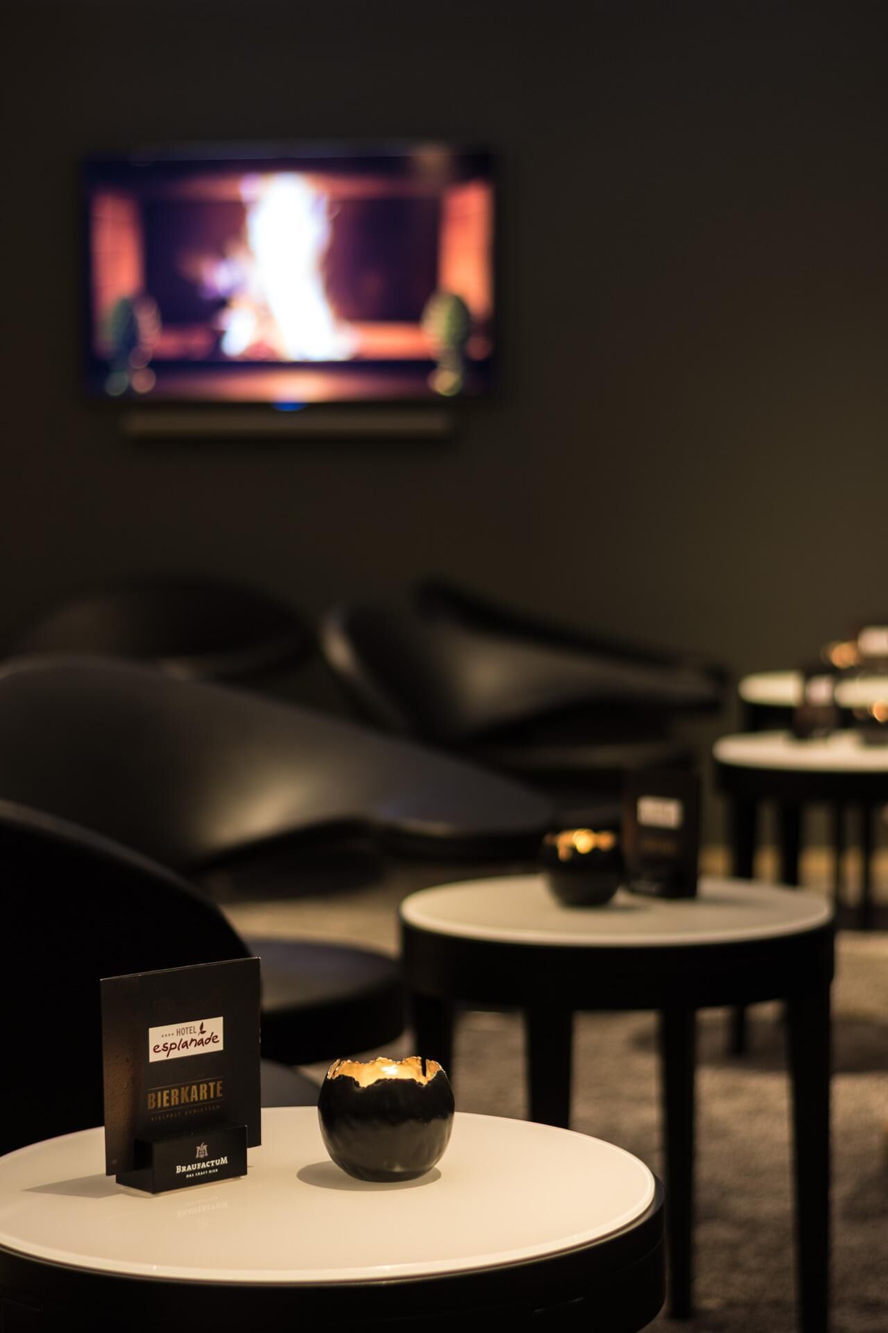 A modern hotel room with a flat-screen TV, black leather seating, and a small table with a candle and a bottle of beer.