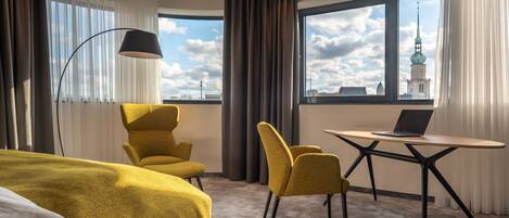 A hotel room with a bed, two chairs, a desk with a laptop, and a large window with a city view.