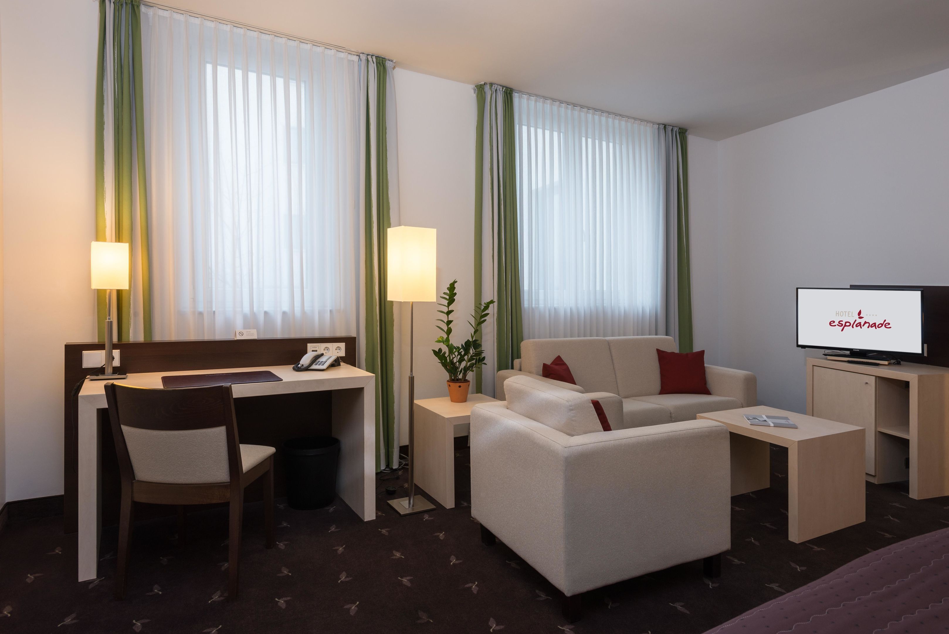 A hotel room with a desk, chair, sofa, television, and a plant.