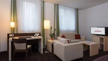 A hotel room with a desk, chair, sofa, television, and a plant.