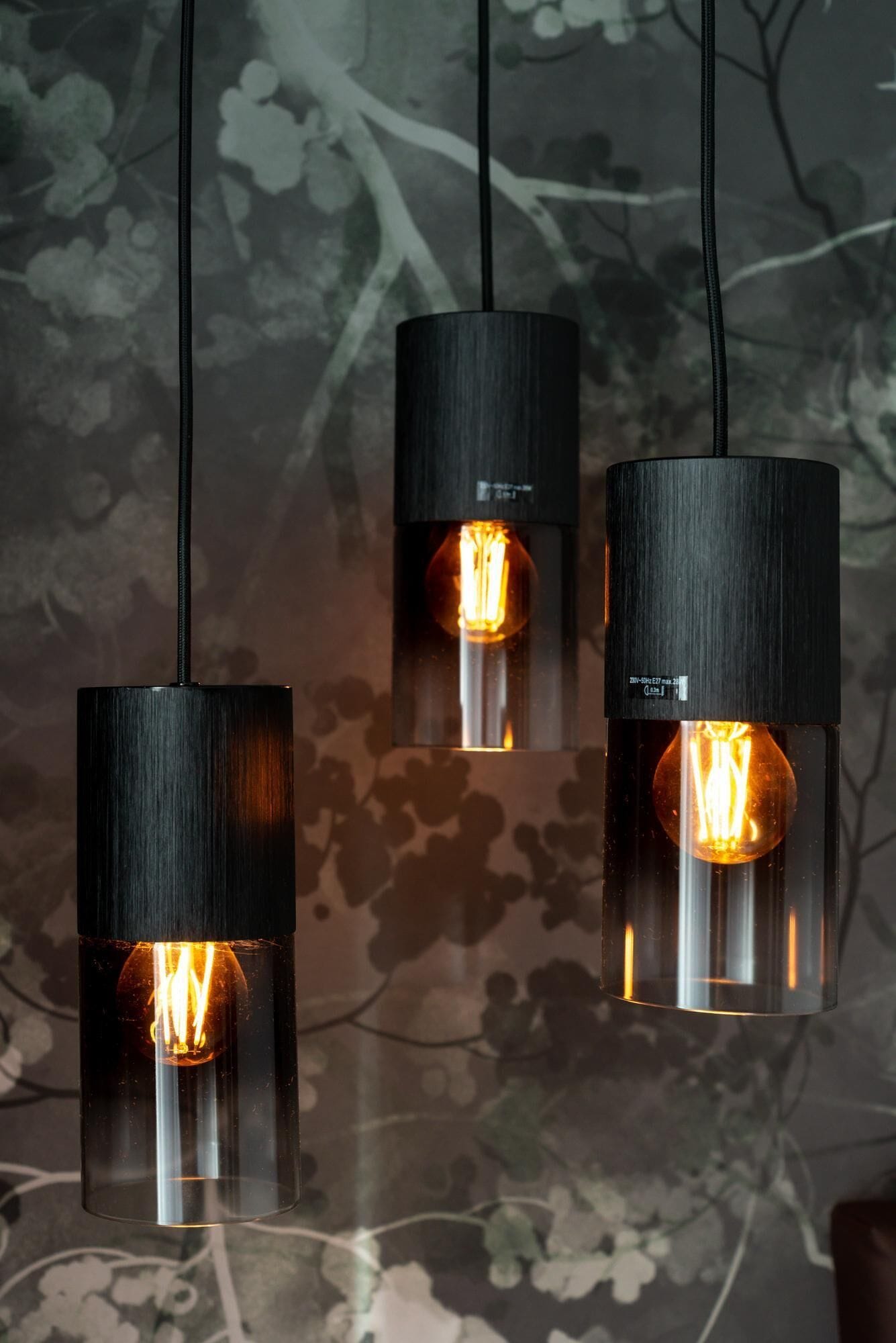 Three pendant lights with exposed filament bulbs, hanging from black cords against a floral-patterned wall.