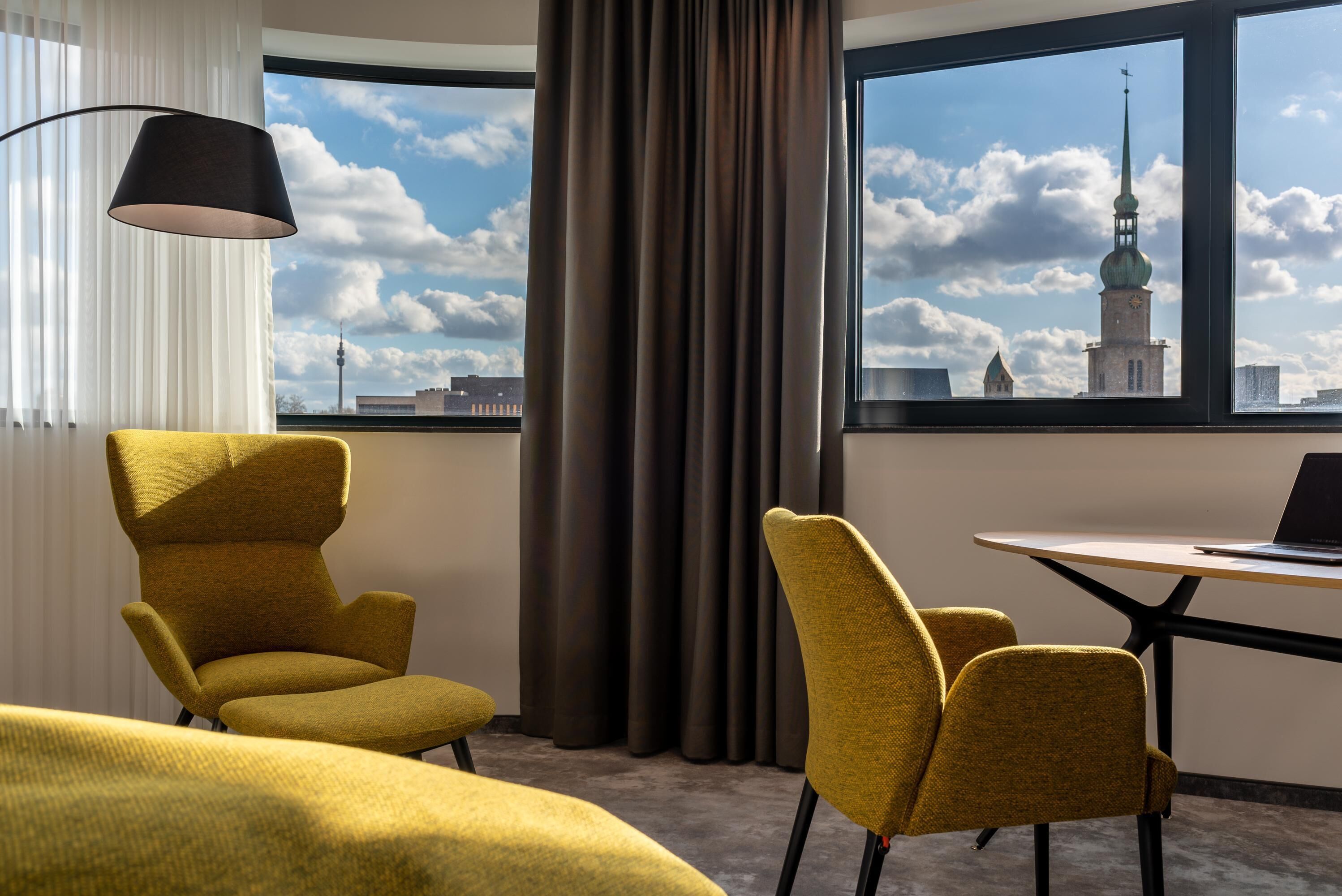 Ein Hotelzimmer mit großem Fenster, zwei gelben Sesseln, einem runden Tisch und einer schwarzen Lampe.