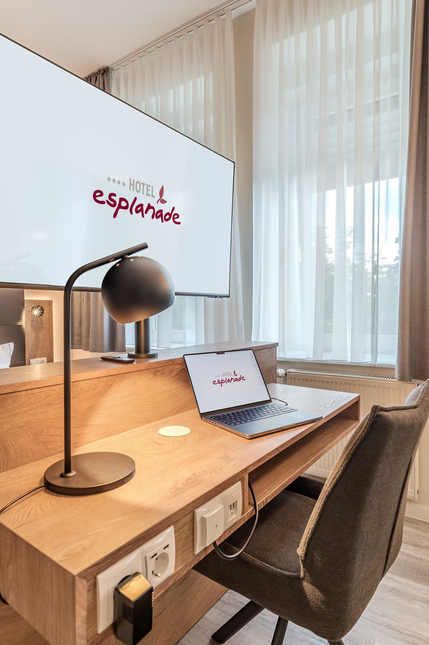 Habitación de hotel con un escritorio de madera, una computadora portátil, una lámpara de escritorio y un amplio ventanal con cortinas translúcidas.