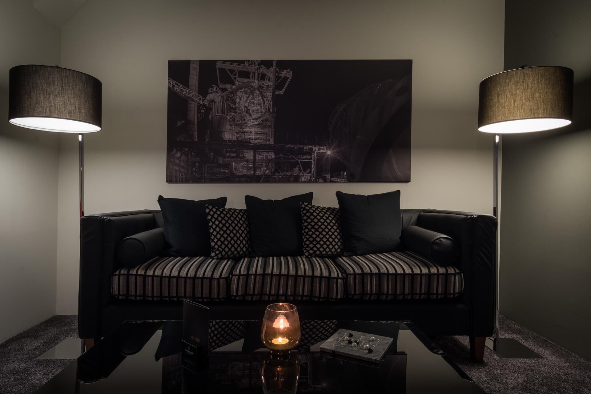 A modern living room with a black sofa, patterned cushions, a glass coffee table, and two floor lamps.