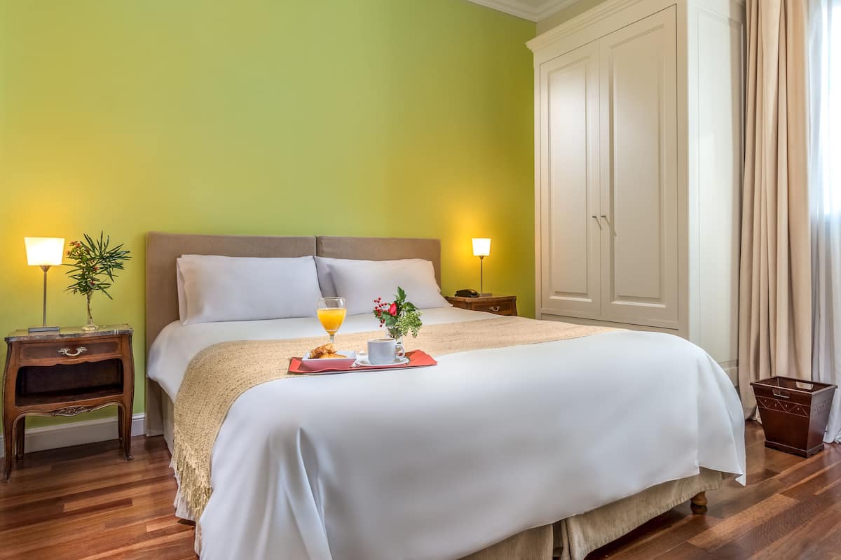 A neatly made bed with white linens, a bedside table with a lamp, a small plant, and a tray with breakfast items.