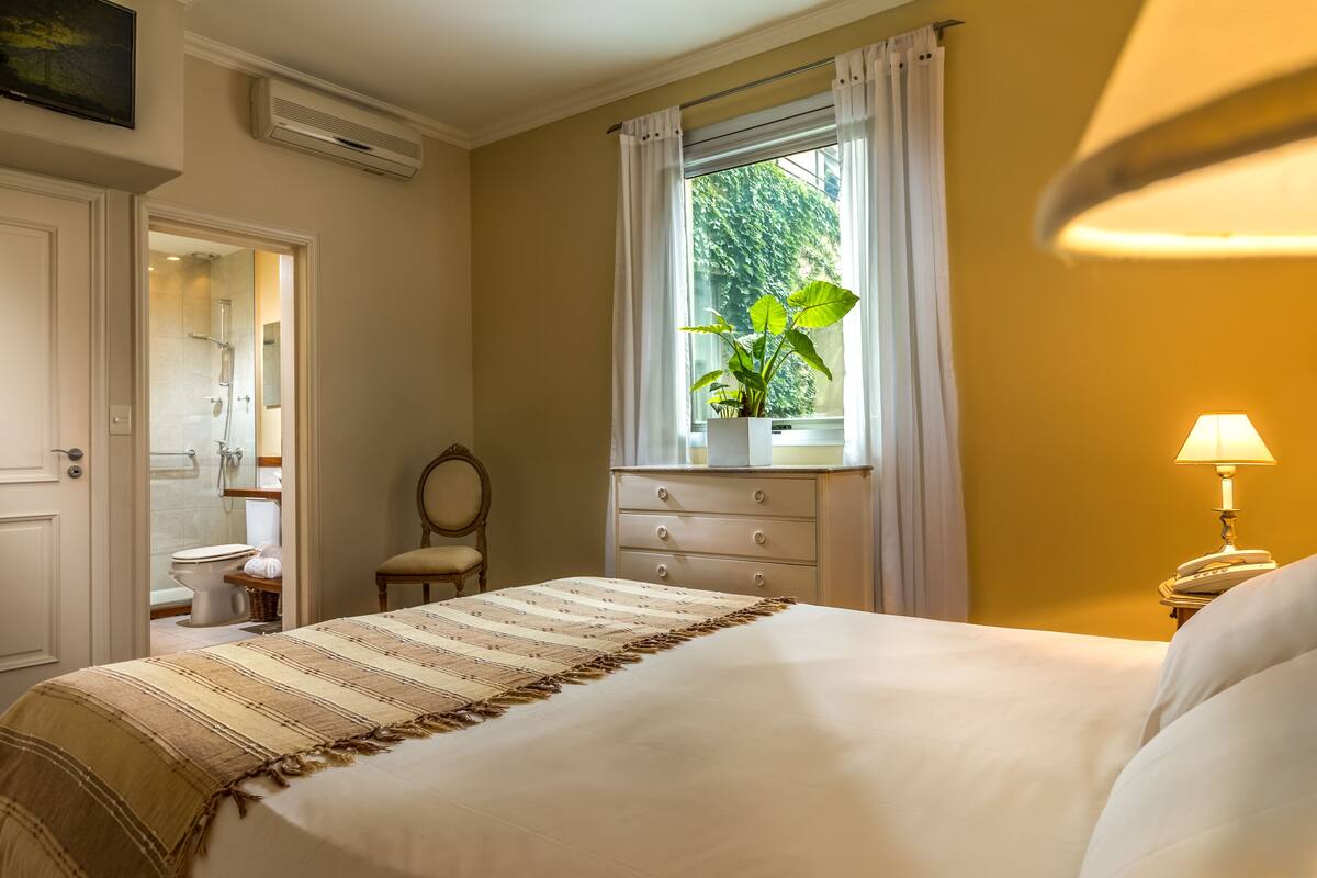 A bedroom with a bed, a window with curtains, a bedside lamp, a chair, and a dresser.