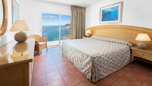 Une chambre d’hôtel avec un lit, un bureau, une chaise, une grande fenêtre donnant sur la mer et un tableau au mur.