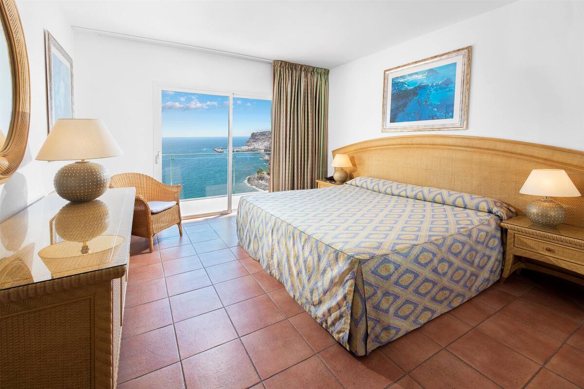 Een hotelkamer met een bed, een bureau, een stoel, een groot raam met uitzicht op zee en een schilderij aan de muur.
