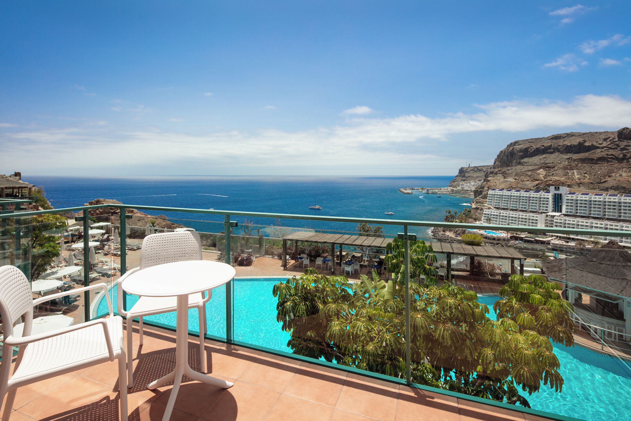 A balcony with a pool, white chairs, and a view of the sea and a resort building.