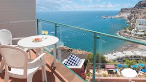Un balcon doté d’une balustrade en verre offrant une vue sur la côte, avec une table, des chaises et un coussin à carreaux.