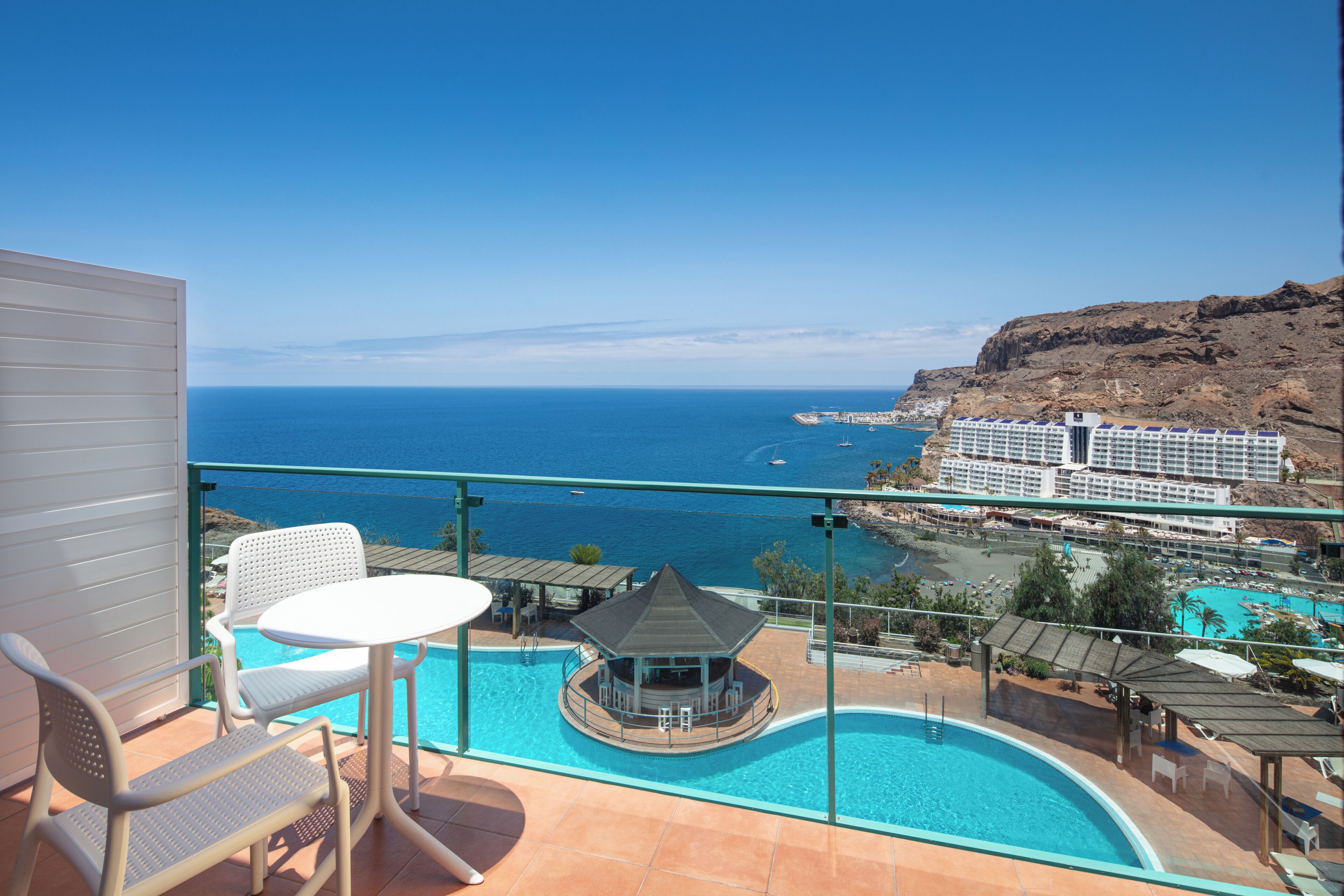 A balcony with a pool, chairs, and a view of the sea and a resort.