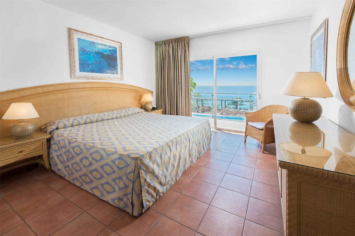 Hotelkamer met een bed, nachtkastje, lamp, stoel, tafel en een groot raam met uitzicht op de oceaan.