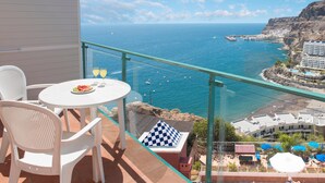 A balcony with a glass railing overlooking a coastal view, featuring a table with chairs and a checkered cushion. -  Princess Mogán & Beach Club All Inclusive (Mogan)