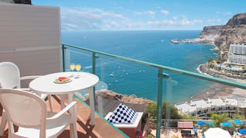 Un balcón con barandilla de vidrio que ofrece vistas al mar, con una mesa, sillas y un cojín a cuadros.