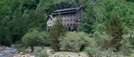 Un edificio in pietra con numerose finestre e un balcone, immerso in una fitta foresta e affiancato da un fiume.