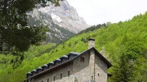 Un edificio de piedra con varias ventanas y un balcón, rodeado de árboles verdes y una montaña al fondo.