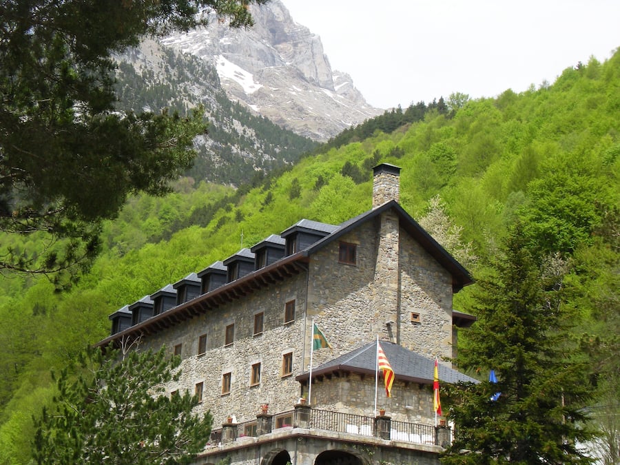 Un edificio de piedra con varias ventanas y un balcón, rodeado de árboles verdes y una montaña al fondo.