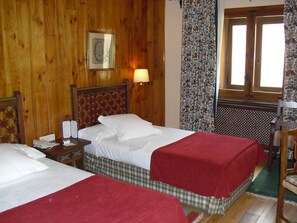 Habitación de hotel con dos camas, paredes de madera, un cuadro enmarcado, una mesita de noche, un teléfono y una ventana con cortinas florales.