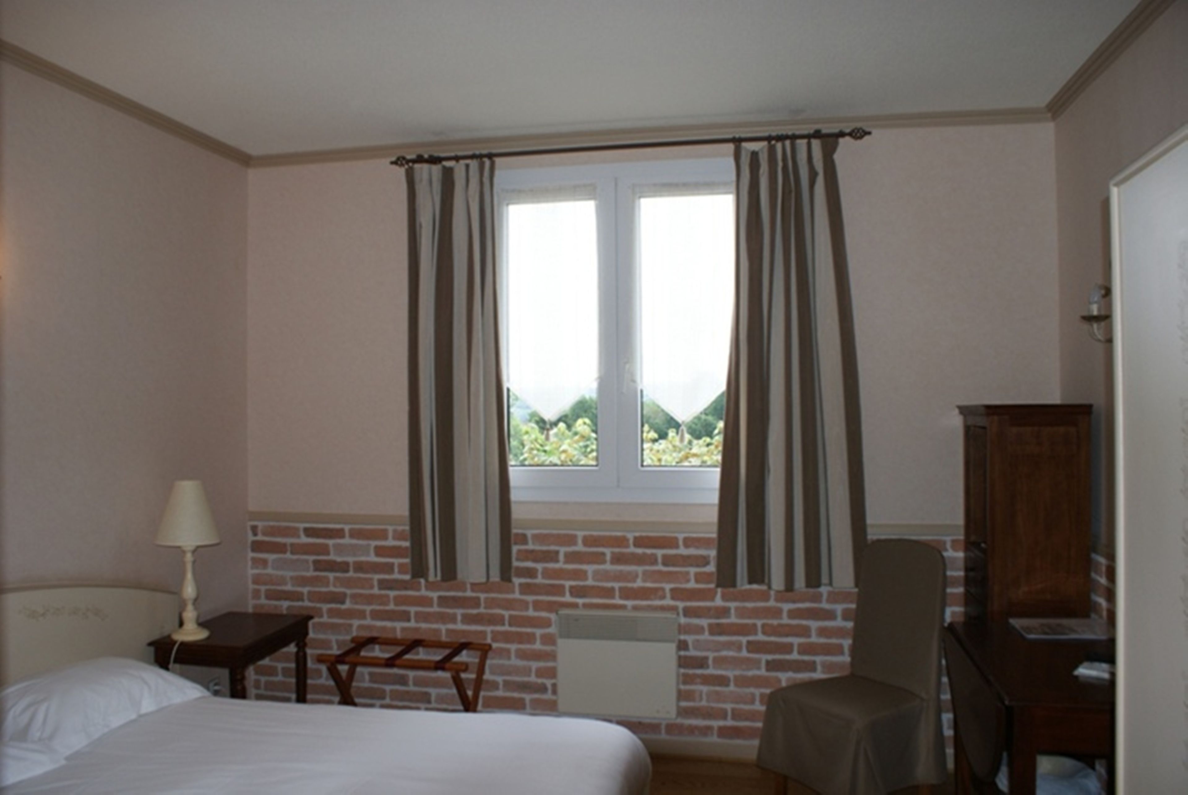 A bedroom with a bed, a nightstand, a chair, a desk, a lamp, and a window with curtains.