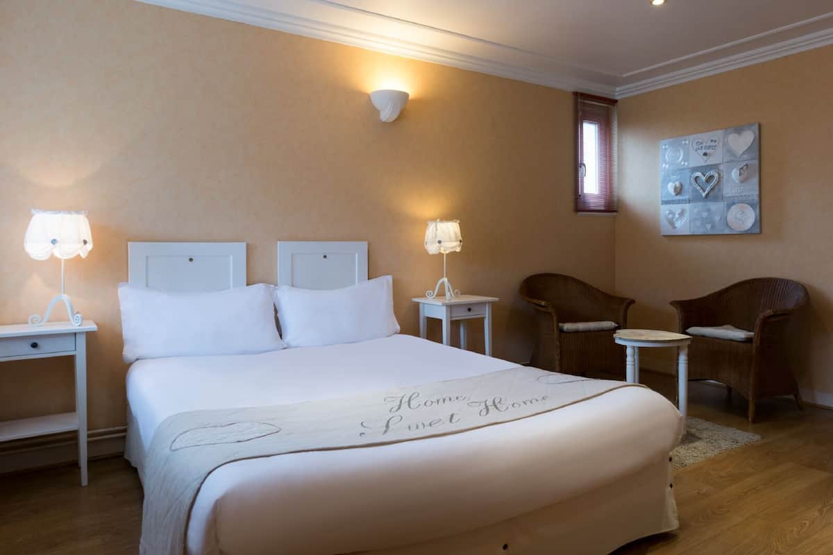 A neatly arranged hotel room with a large bed, two bedside tables with lamps, a small round table, two wicker chairs, and a decorative wall piece.