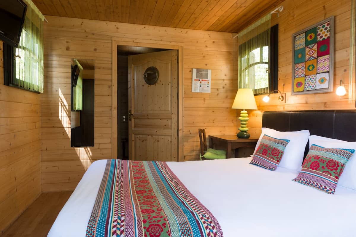 A wooden cabin room with a bed, a table with a lamp, and a door.