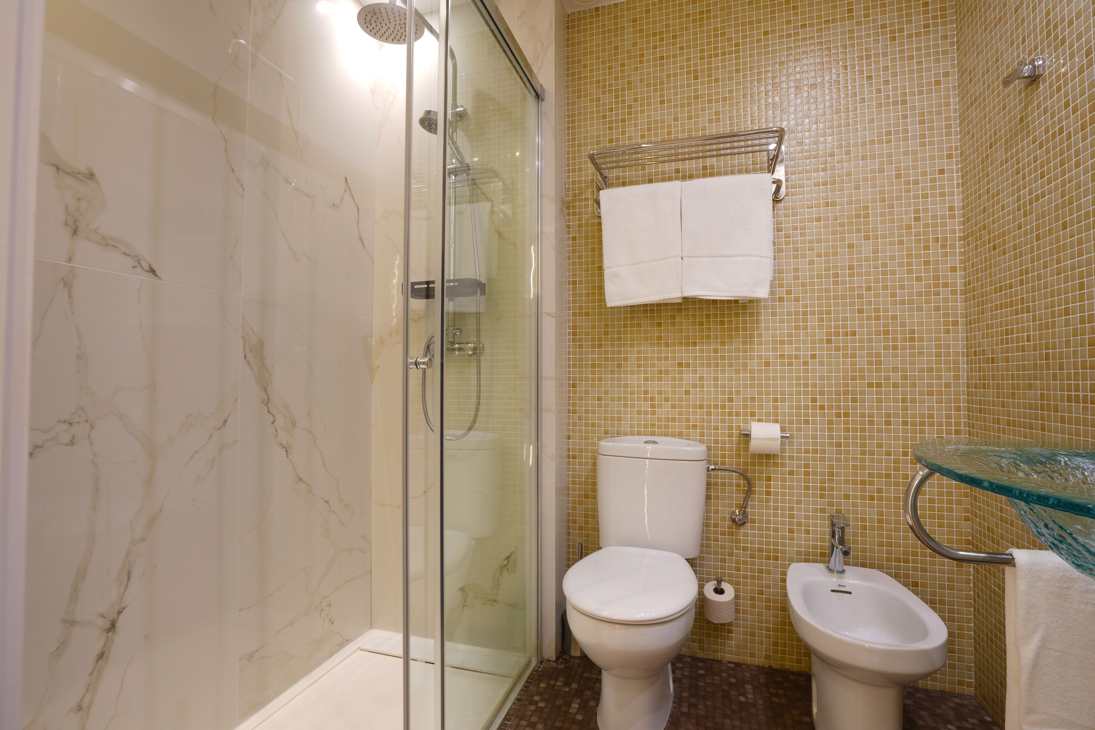 A bathroom with a glass shower enclosure, a toilet, a bidet, and a glass sink.