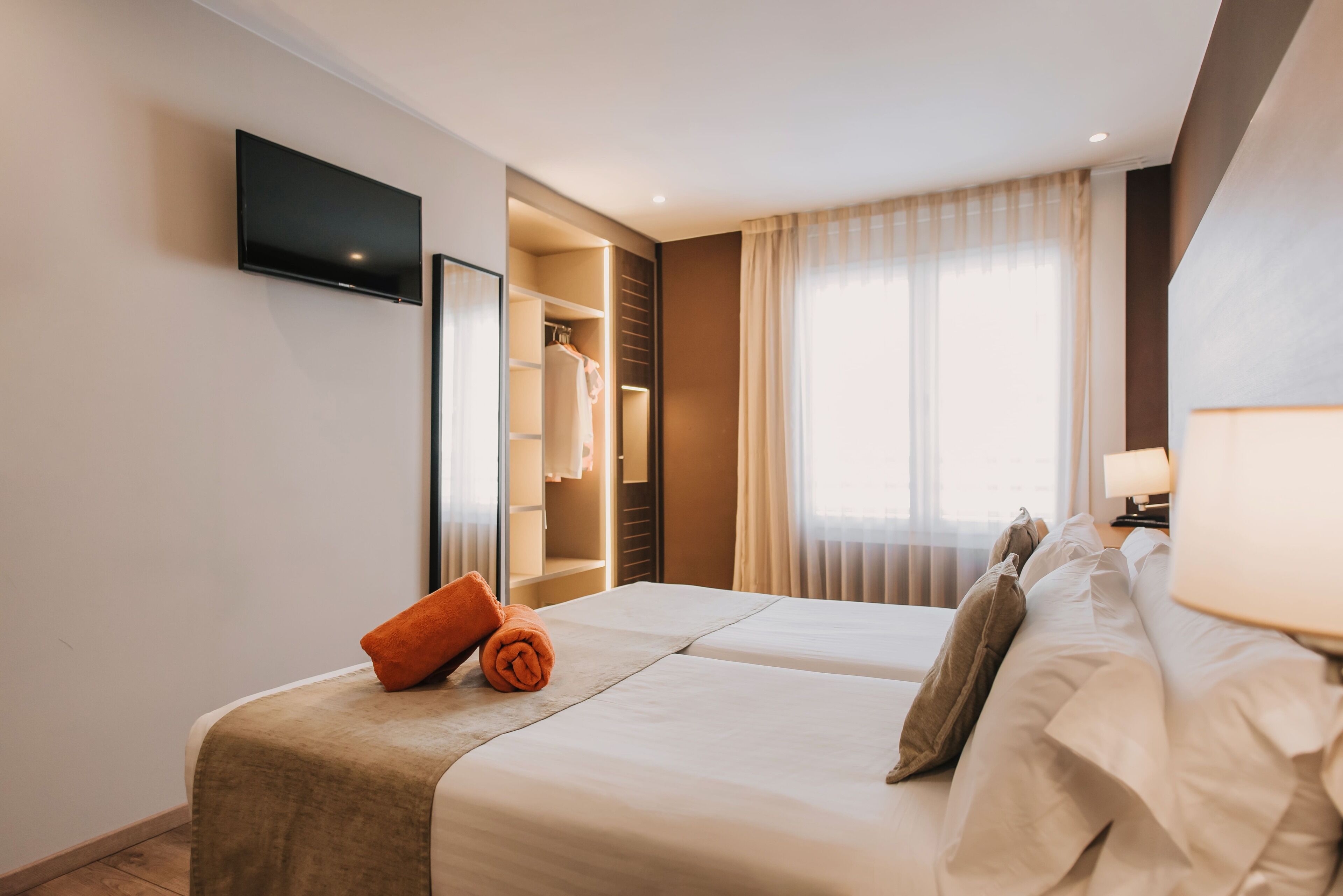 Une chambre d’hôtel avec un lit, un téléviseur fixé au mur, un dressing et une fenêtre avec des rideaux.
