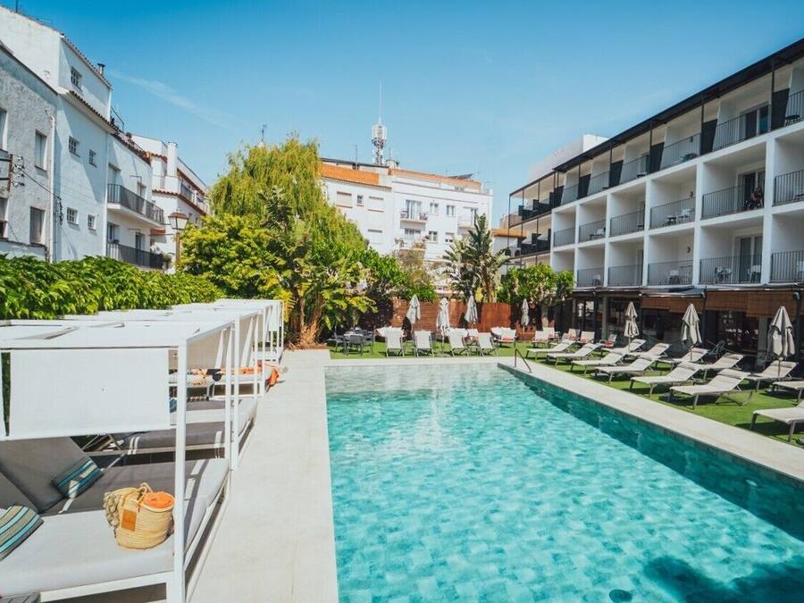 Piscina con sillones de descanso y sombrillas, rodeada de edificios y vegetación.