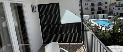 Een balkon met een witte tafel en stoelen, een donker privacy scherm, en uitzicht op een zwembad en gebouwen in de verte.