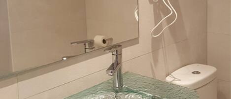 A bathroom with a glass sink, a hairdryer mounted on the wall, and a toilet.