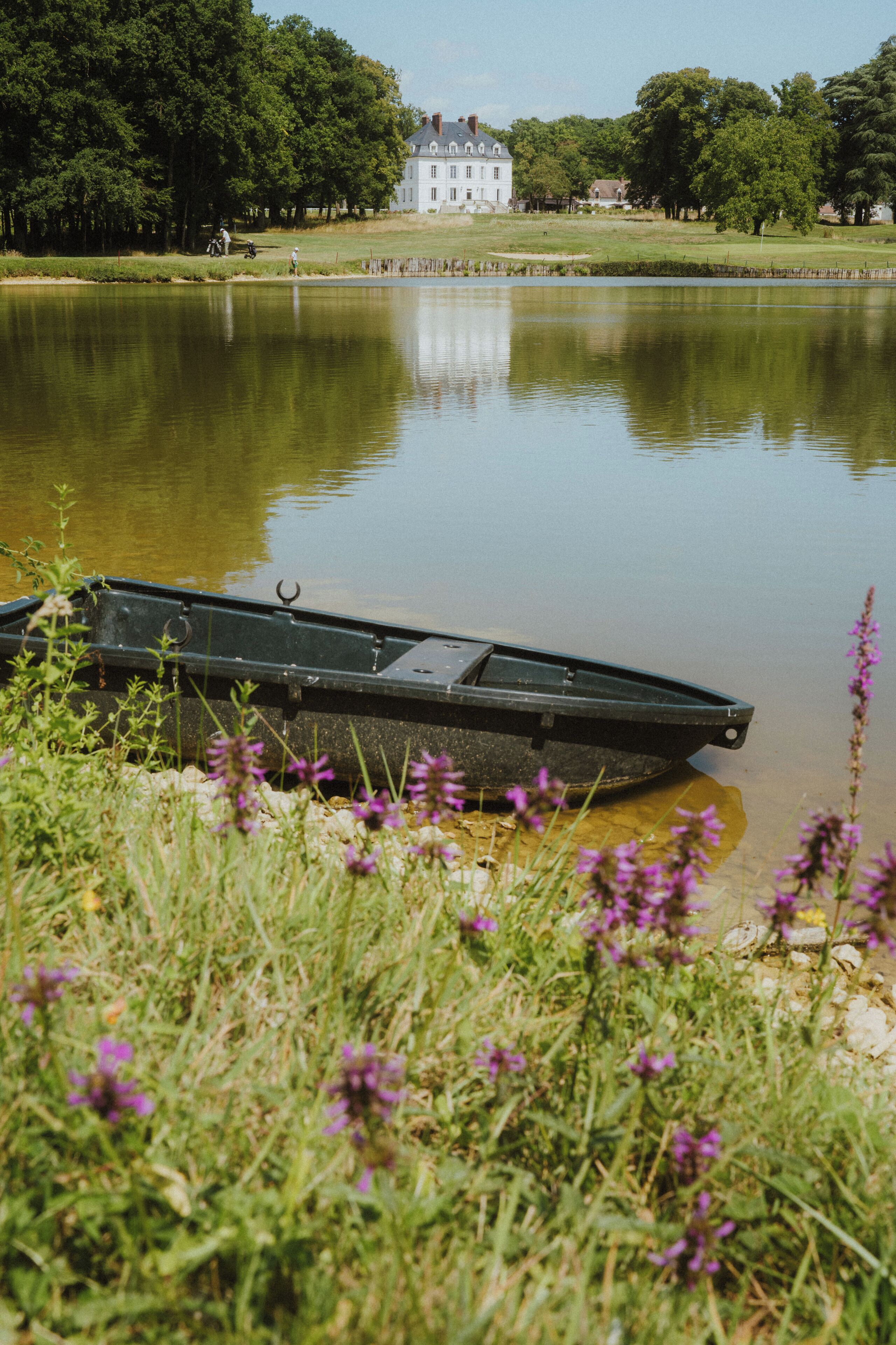 Un lago tranquilo con un bote, una casa grande al fondo y abundante vegetación alrededor.