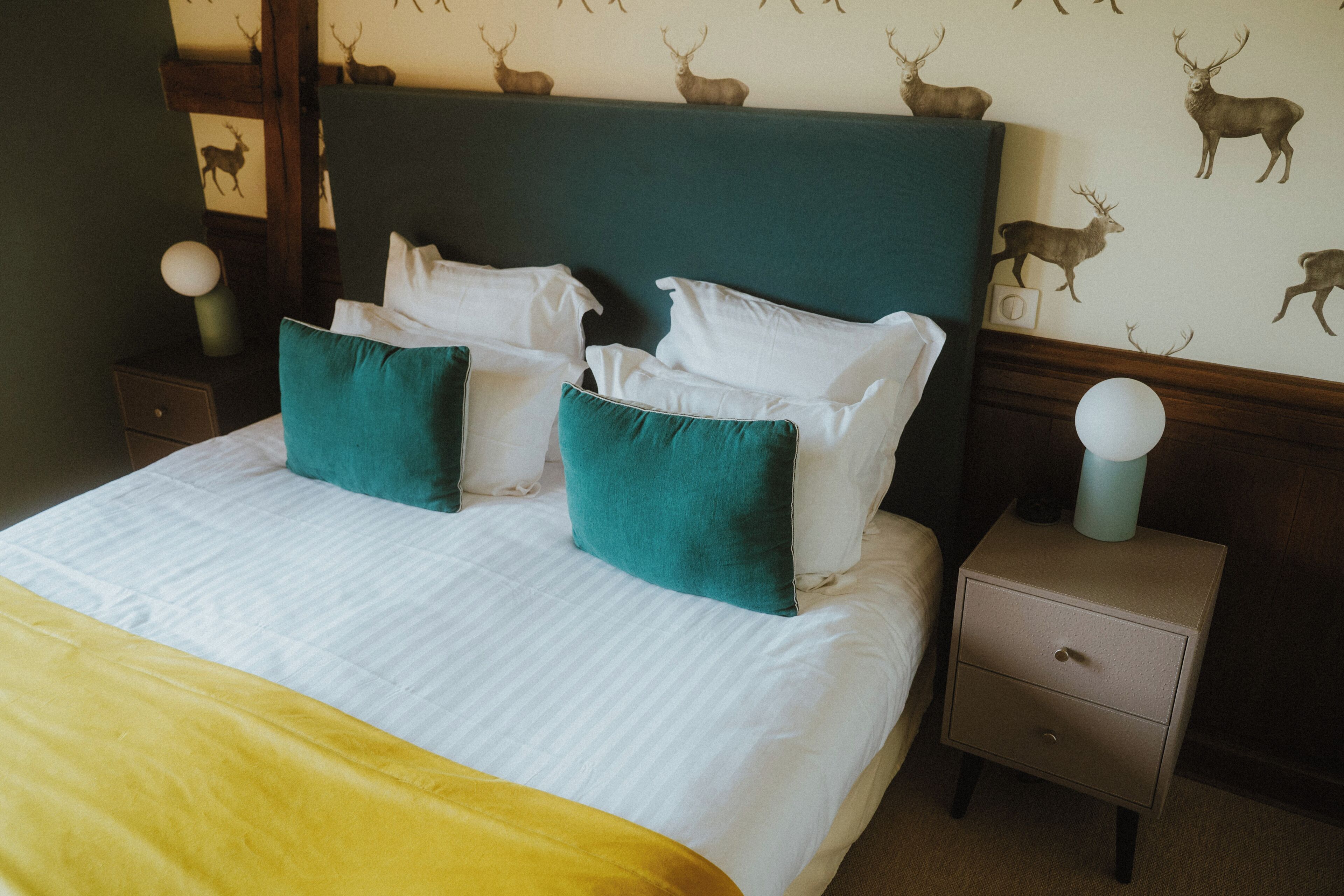 A neatly made bed with white linens and teal pillows, a wooden headboard, a nightstand with a lamp, and a wall with deer decals.