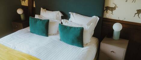 A neatly made bed with white linens and teal pillows, a wooden headboard, a nightstand with a lamp, and a wall with deer decals.