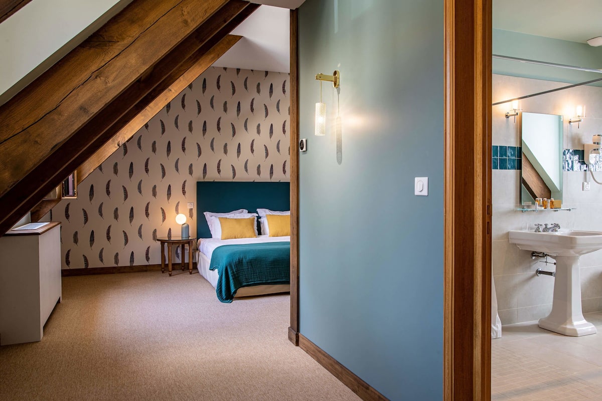 A room with a bed, a bedside table, a wall-mounted light, a mirror, and a bathroom with a sink and tiles.