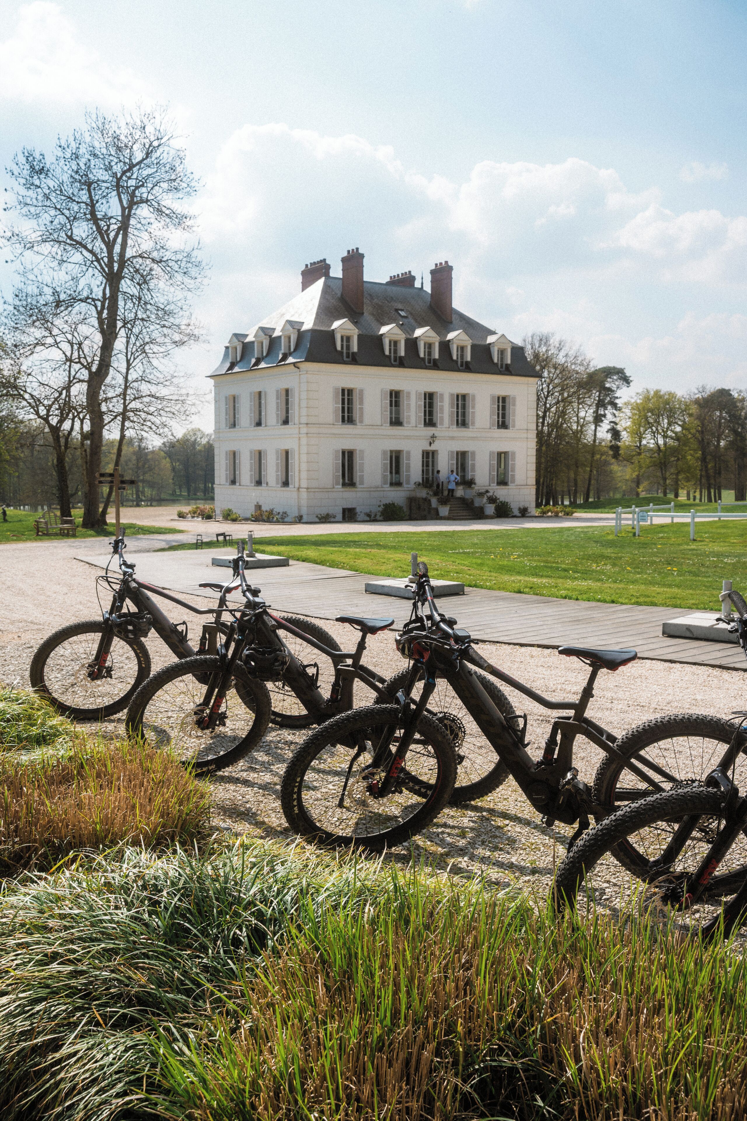 Un grand bâtiment à la façade symétrique, une zone pavée où sont garés des vélos et un espace verdoyant en premier plan.