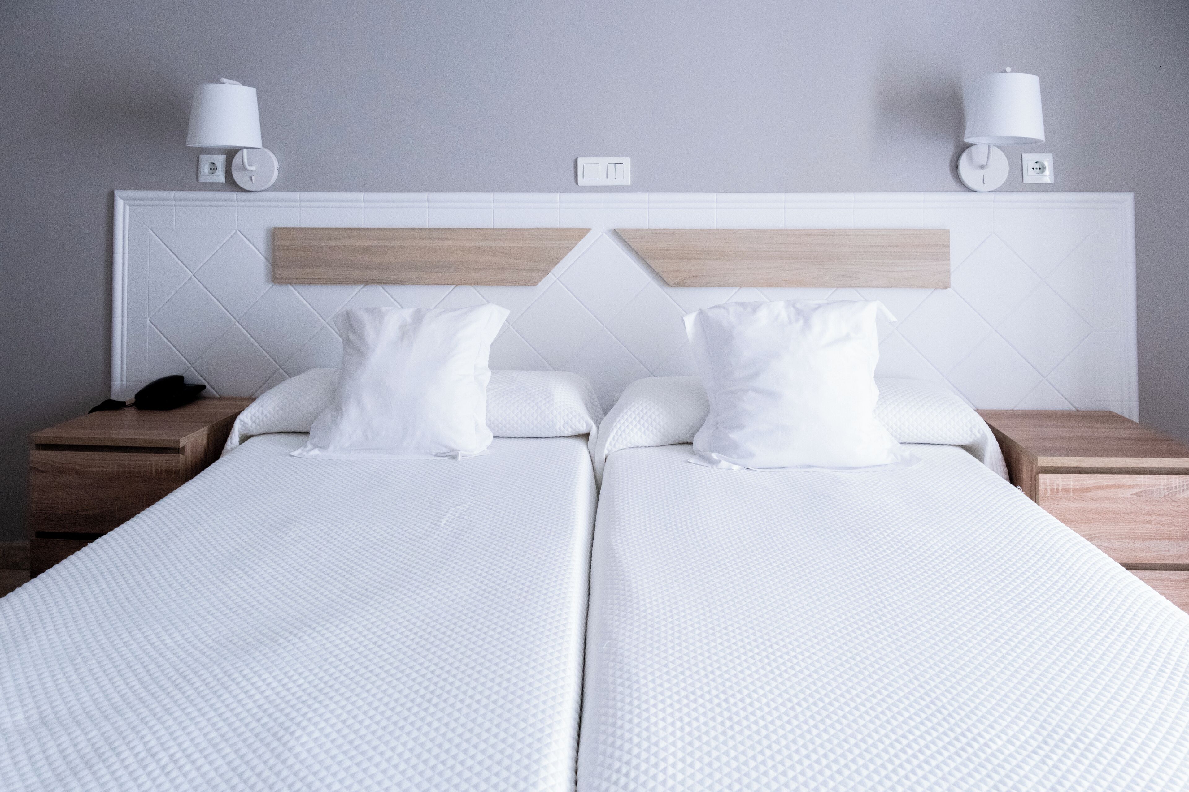 Habitación de hotel con dos camas, ropa de cama blanca, cabeceros de madera, mesitas de noche y lámparas de pared.