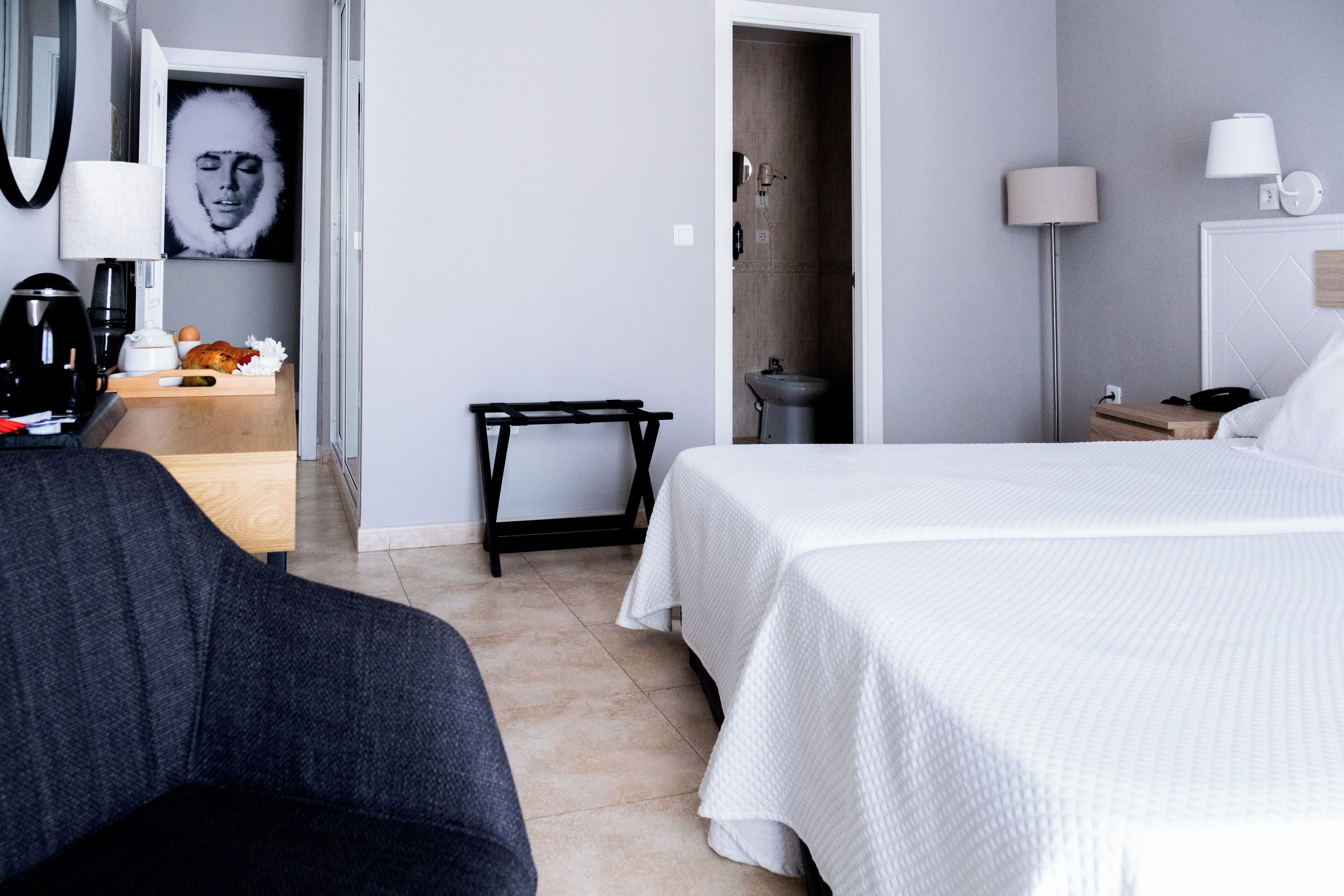A hotel room with a bed, a desk with a lamp, a chair, a small table with a cup and a tissue box, and a door leading to a bathroom.