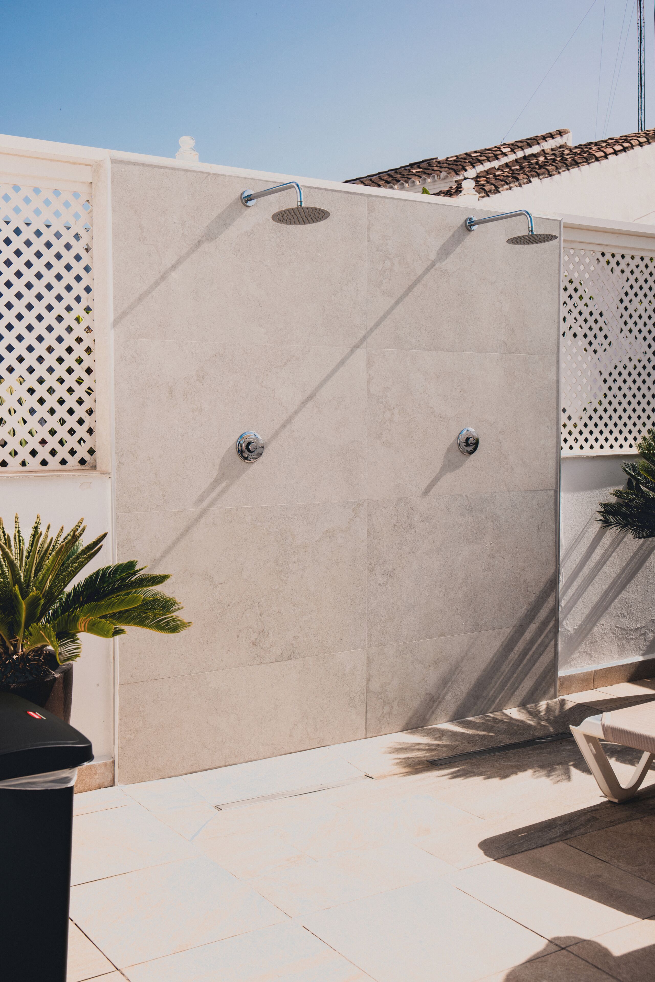 A modern outdoor shower with a tiled floor, a white wall, and a decorative fence.