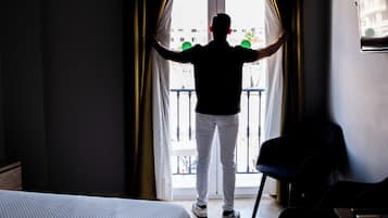Ein Hotelzimmer mit einem Bett, einem Schreibtisch und einem Stuhl. Ein Fenster mit Vorhängen ist zu sehen, und dahinter ist ein Balkon erkennbar.