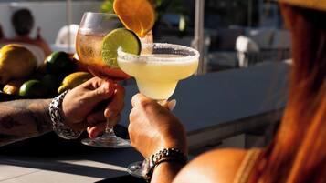 Two people holding drinks with lime and orange slices, a plate with more fruit, and a large umbrella in a sunny outdoor setting.
