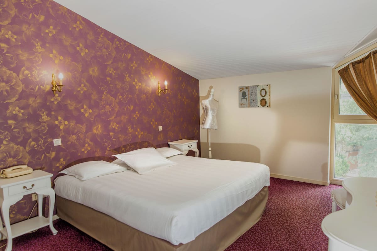 Une chambre d’hôtel avec un grand lit, une table de chevet avec un téléphone, une lampe fixée au mur et une fenêtre avec des rideaux.