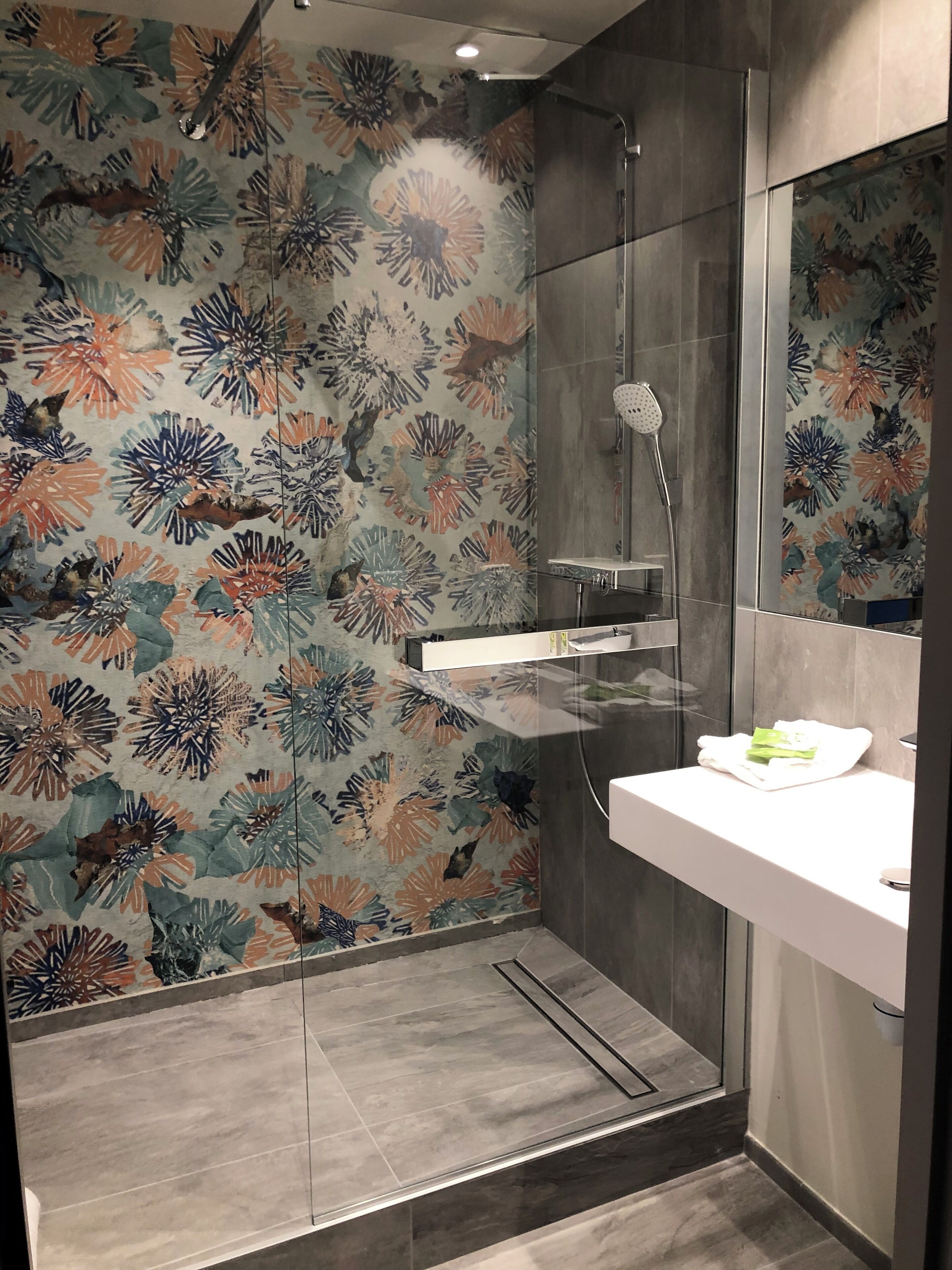 A modern bathroom with a shower, a glass-enclosed vanity, and a floral-patterned wall.