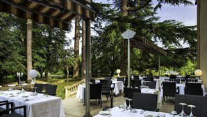 Een buitenruimte met eethoek, tafels voor twee personen, omringd door bomen en overdekt met een pergola.