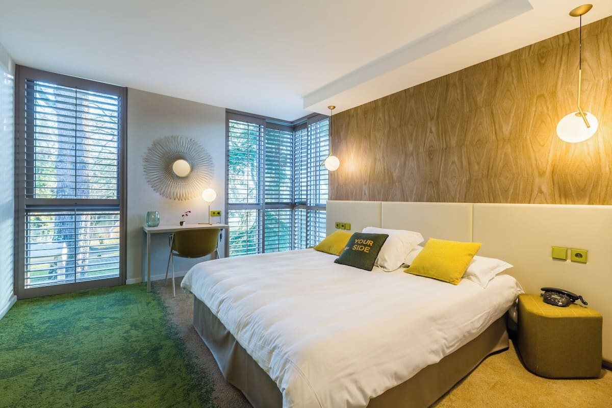 Une chambre moderne avec un grand lit, un bureau et une chaise. On y trouve plusieurs fenêtres équipées de stores, un grand miroir rond et un luminaire fixé au mur.
