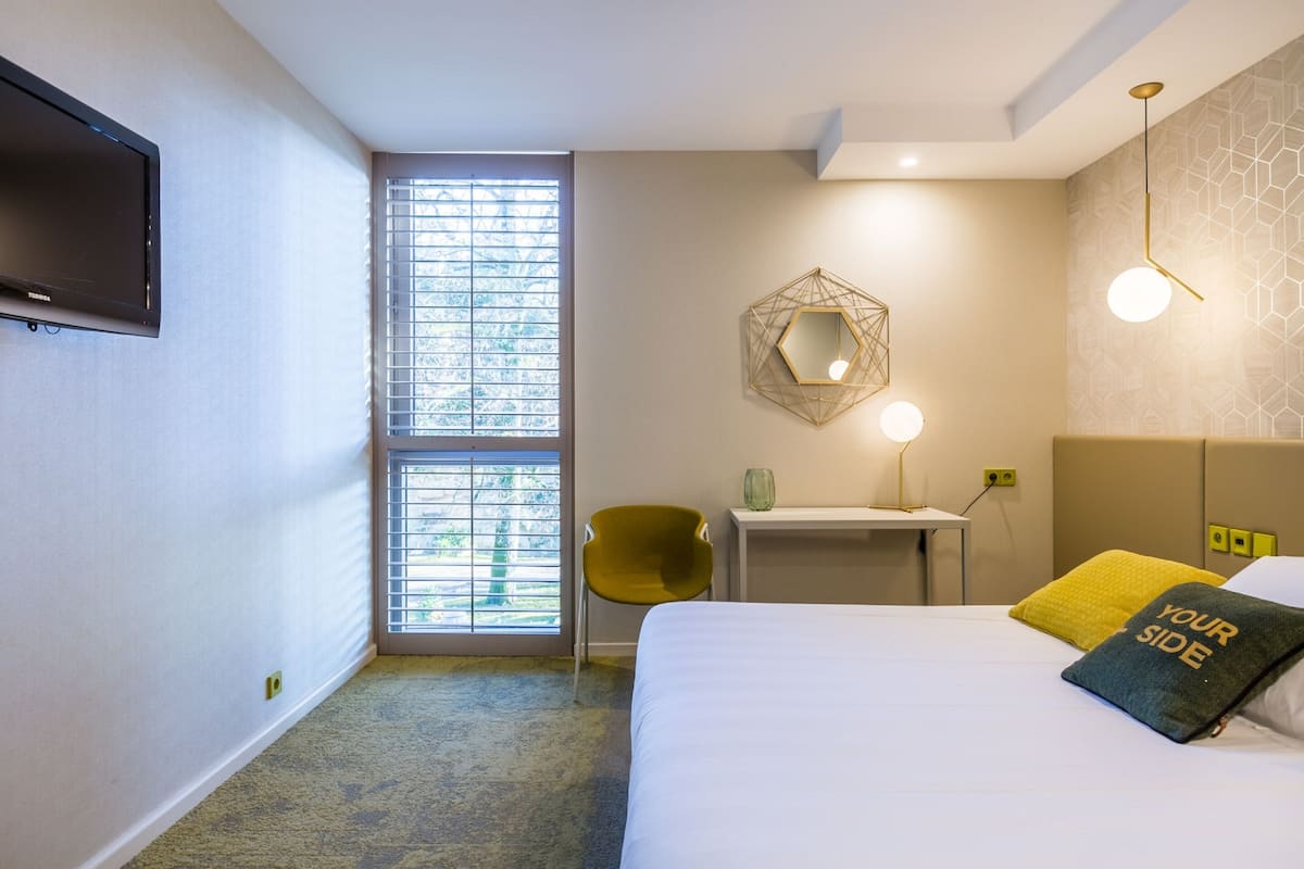 Une chambre d’hôtel équipée d’un lit, d’un bureau, d’une chaise, d’une télévision et d’une fenêtre avec des stores.