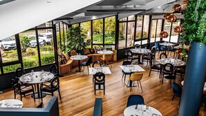 Een restaurant met ronde tafels, stoelen en uitzicht op geparkeerde auto's door grote ramen.